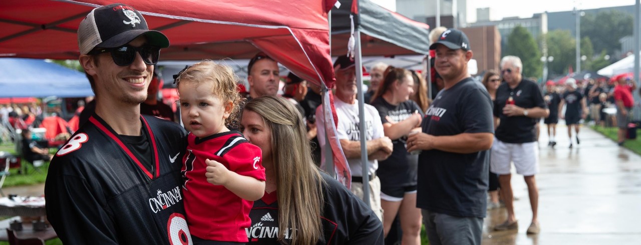 man wearing a UC football jersey, holding a toddler during tailgating festivities before a home football game